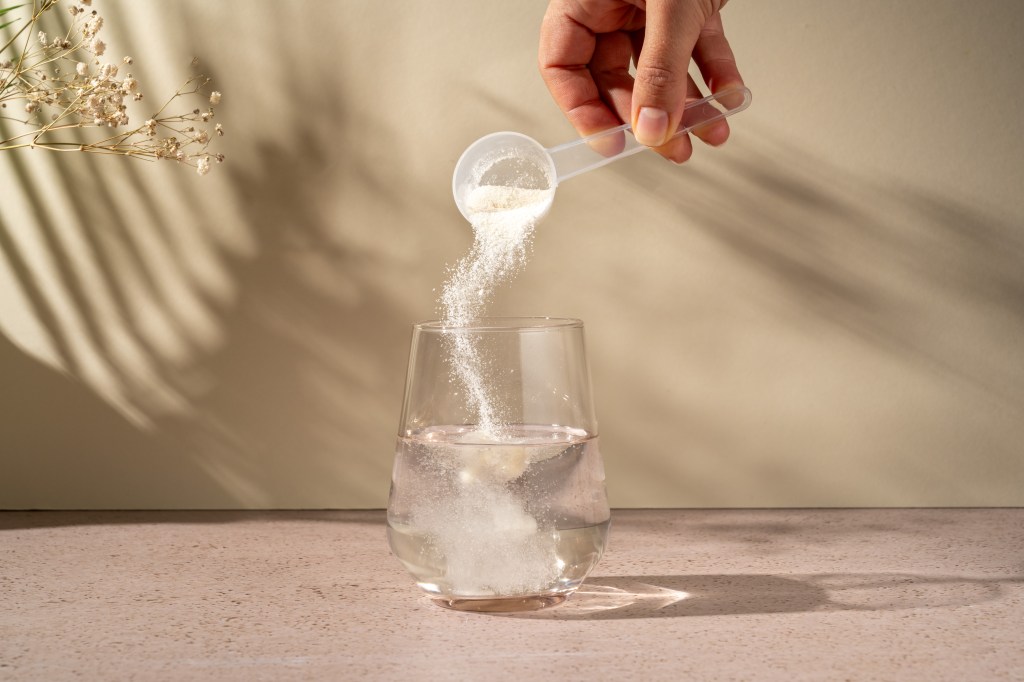 A hand pouring collagen protein powder from a scoop into a glass of water.