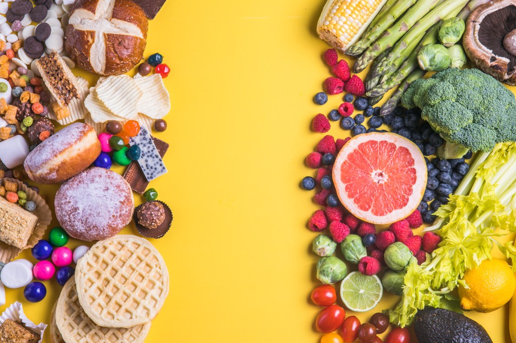 A split overhead view of unhealthy and healthy foods on a yellow background. The left side has fast food, sweets, and snacks, while the right side contains fruits and vegetables.