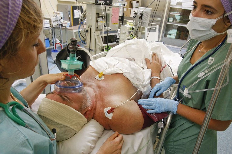 A person with two attending health-care workers is on a surgical bed and is being anaesthetized for brain surgery.