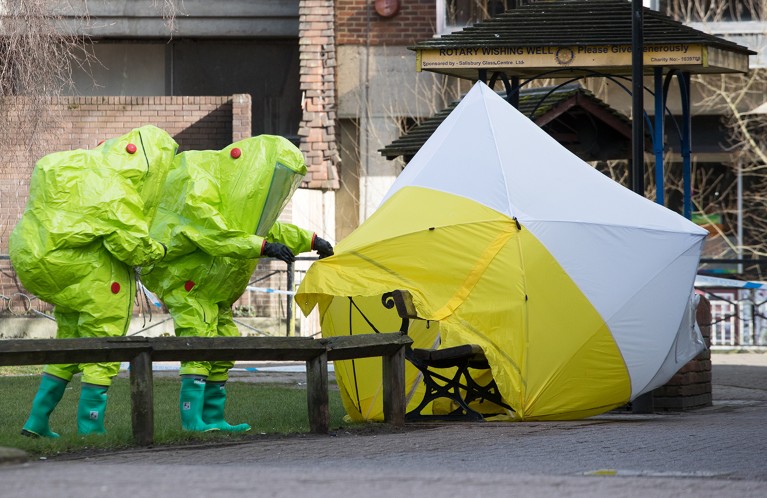 Two people in full hazmat suits approach a park bench partially covered by a forensic tent.