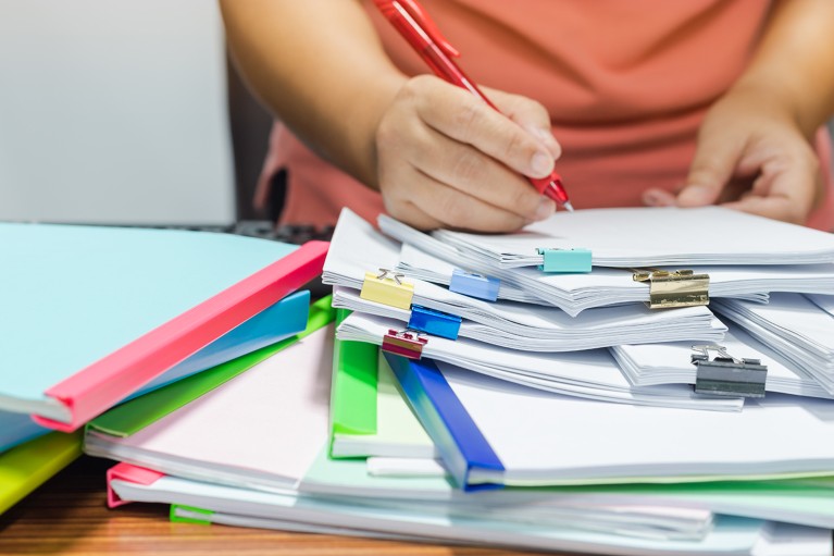 Closeup of a hand holding a red pen checking papers on a stack of reports.