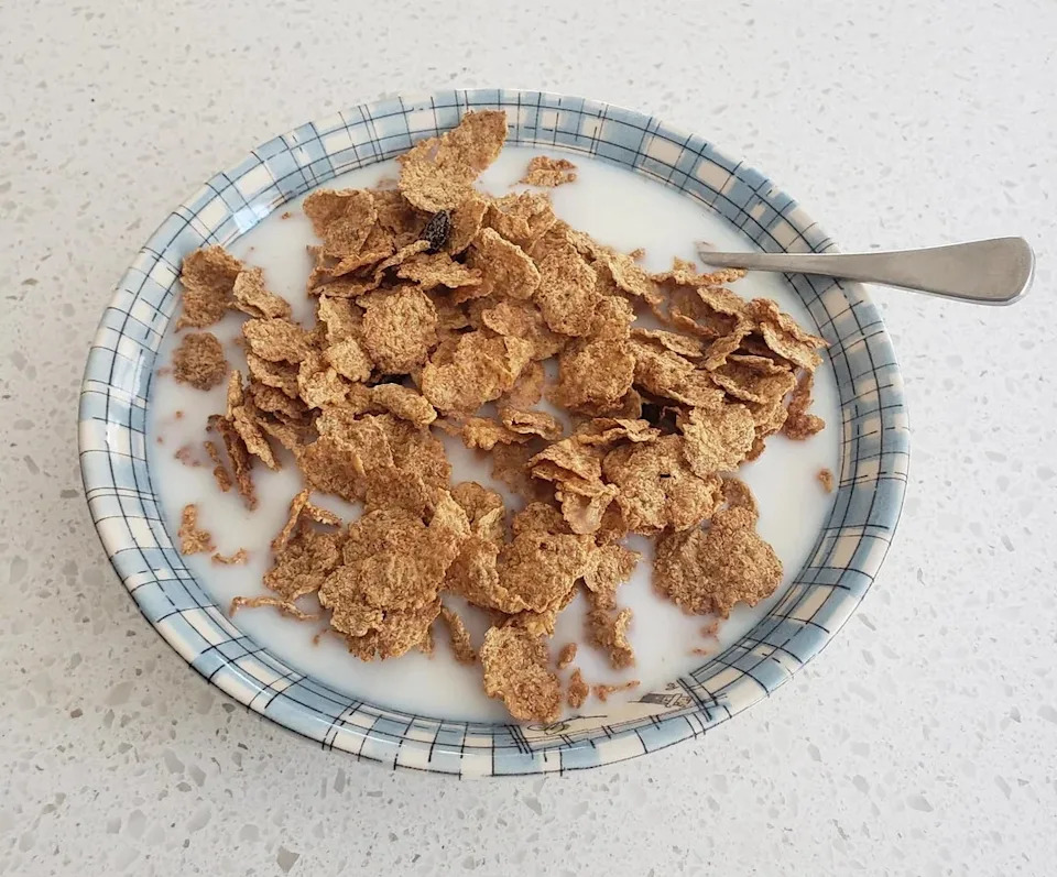 A bowl of bran flakes with milk on a checkered plate, with a spoon resting inside