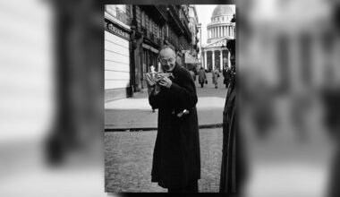 A man in a dark coat smiles while holding a cat on a city street. In the background, pedestrians walk near classical buildings, including a domed structure. The scene appears to be in an urban European setting.