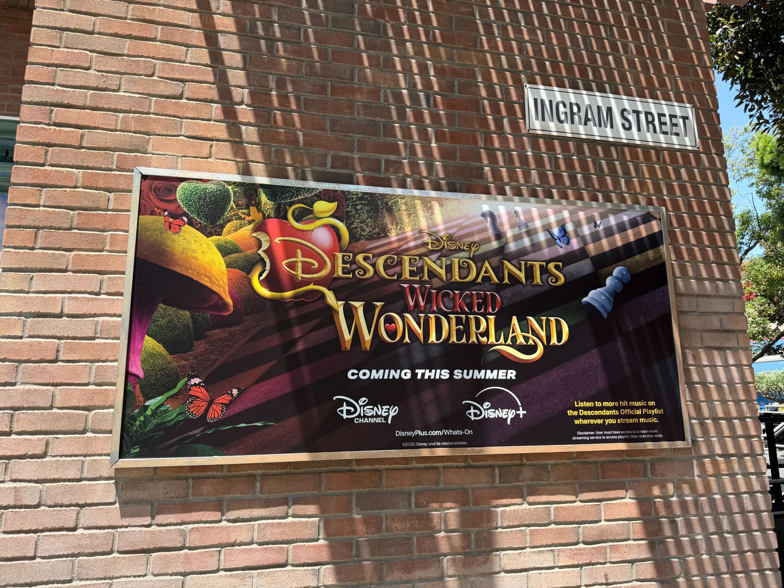 A billboard for the movie is installed on a brick wall nearby. It informs guests that the movie will be coming this summer on Disney Channel and Disney+ and there is an official Descendants playlist that can be streamed.