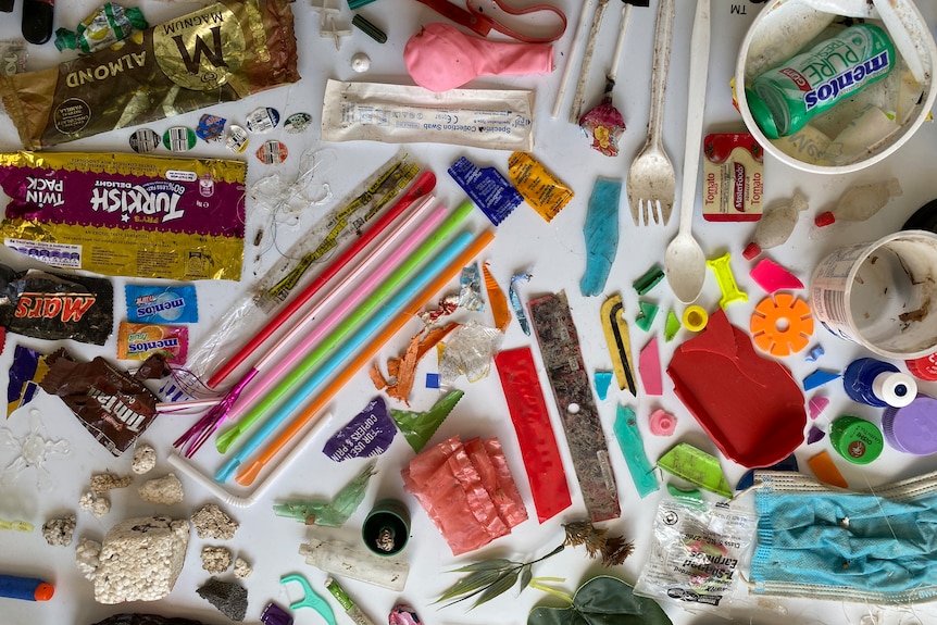 Plastic utensils, brightly coloured wrappers and straws and other odd shapes of plastic spaced out on white background.