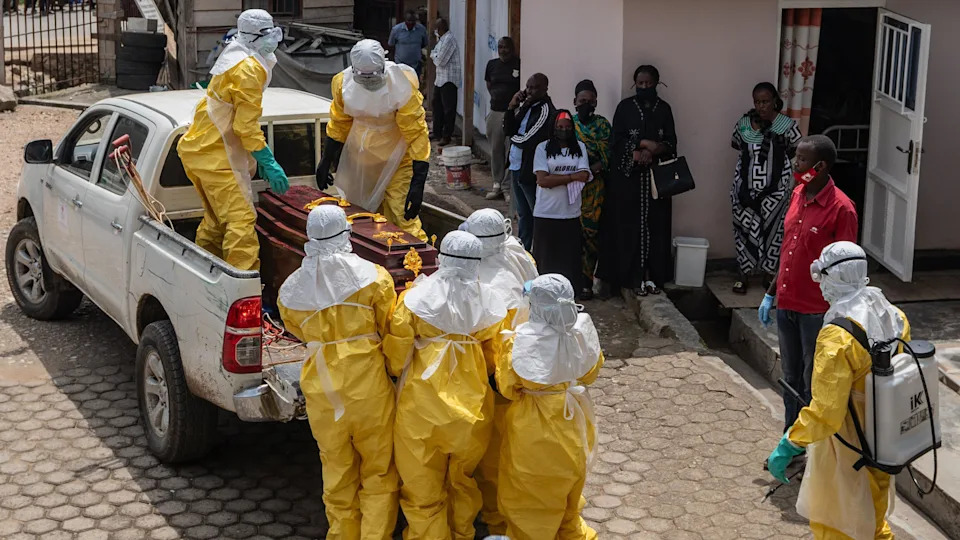 Health workers wearing full personal protective equipment (PPE) lift a coffin of a victim on to the back of a pick-up truck in Bunia. Some people with masks watch them outside a hospital.