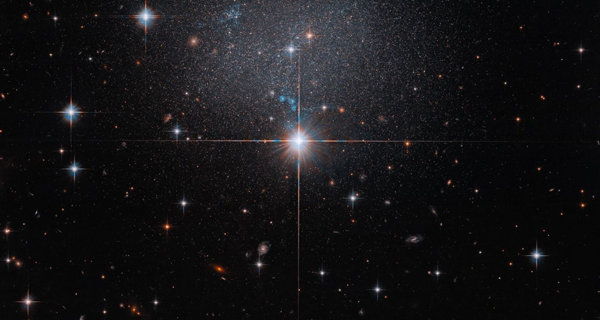 A star in the foreground shines brightly with four diffraction spikes. Above this star is a diffuse and faint dwarf irregular galaxy. The galaxy itself has bright blue and red stars.