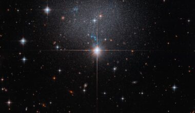 A star in the foreground shines brightly with four diffraction spikes. Above this star is a diffuse and faint dwarf irregular galaxy. The galaxy itself has bright blue and red stars.