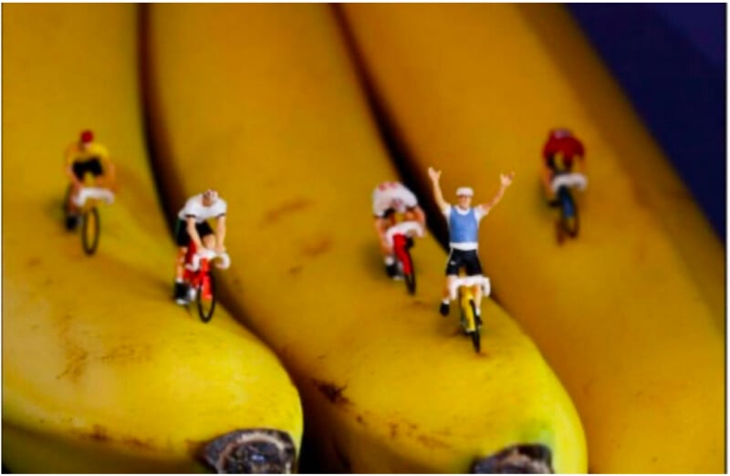 Five miniature figurines riding bicycles are positioned on yellow bananas, resembling a playful bike race on a bright, fruit-themed track. The cyclist in front raises both arms in celebration.
