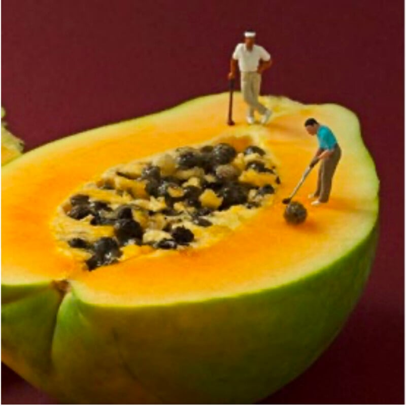 Two miniature figurines appear to play golf on the seeds of a halved papaya, as if the fruit’s seeds are a golf course, against a dark maroon background.