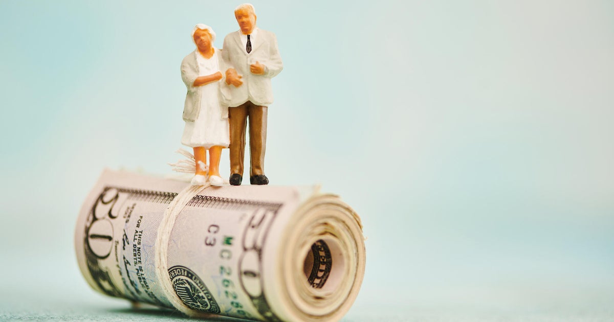 Miniature figures of elderly couple standing on top of a wad of fifty dollar bills