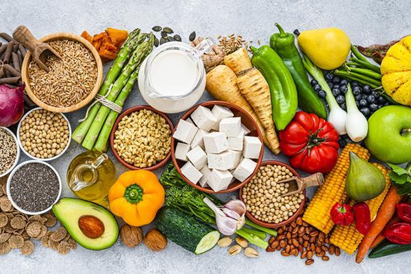 Overhead view of healthy organic foods; the artfully arranged display includes tofu, carrots, broccoli, whole-grain pasta, chia seeds, quinoa, berries, legumes, olive oil, avocado, corn, cucumber, onion, lettuce, tomato, garlic, bell pepper, radish, and more.