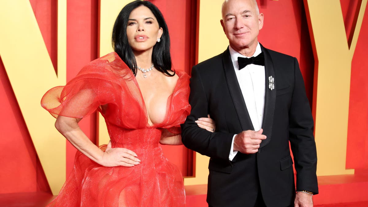 Lauren Sánchez wearing a red gown standing with Jeff Bezos in a tuxedo on a red carpet