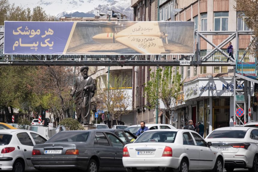 Vehicles stop beneath a banner depicting an Iran-made unmanned aerial vehicle (UAV), the Shahed-136, alongside a Persian script reading 'Each missile is one message,' in Tehran, Iran, on March 30.