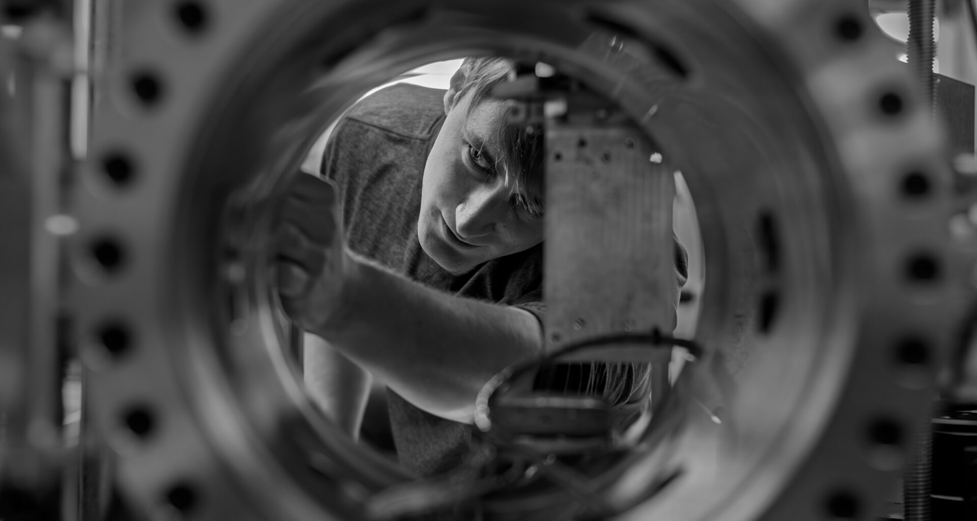 A black-and-white photo of a cryogenic engineer viewed through a circular opening of a large metallic chamber with evenly spaced bolts along its rim. Inside, a square component is mounted with wires connected to it. The man viewed through the porthole appears to be adjusting settings for the machine, which can mimic the extreme cold of the lunar night.
