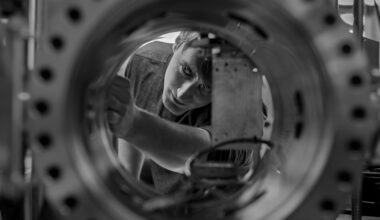 A black-and-white photo of a cryogenic engineer viewed through a circular opening of a large metallic chamber with evenly spaced bolts along its rim. Inside, a square component is mounted with wires connected to it. The man viewed through the porthole appears to be adjusting settings for the machine, which can mimic the extreme cold of the lunar night.