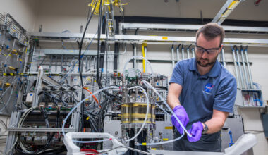 A man wearing a blue NASA polo, purple gloves, and safety glasses adjusts a small tube connected to a silver and yellow cylinder inside the Fuel Cell Testing Laboratory. Behind him are numerous other tubes, wires, and sensors connected to a large system and the lab’s wall.