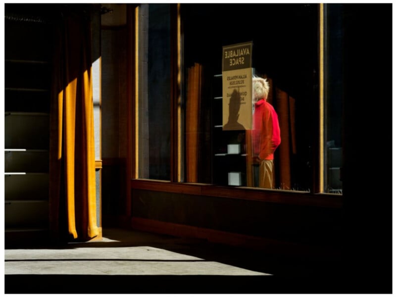 A person wearing a bright red jacket stands in the shadowy interior of a storefront, partially visible through a window with an "AVAILABLE SPACE" sign taped to it. Light and shadow create dramatic contrasts in the scene.