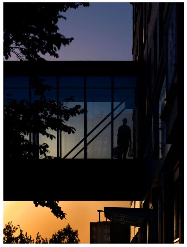 A silhouette of a person carrying bags walks across a glass skybridge at sunset, with a clear evening sky and tree branches framing the scene.