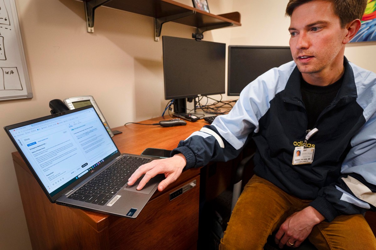 A person sits at a desk pointing to a laptop screen displaying a document, with two monitors and office items in the background.