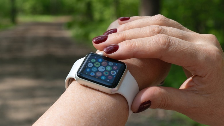 An individual with dark red nail polish adjusts their Apple Watch while they are on a trail surrounded by foliage
