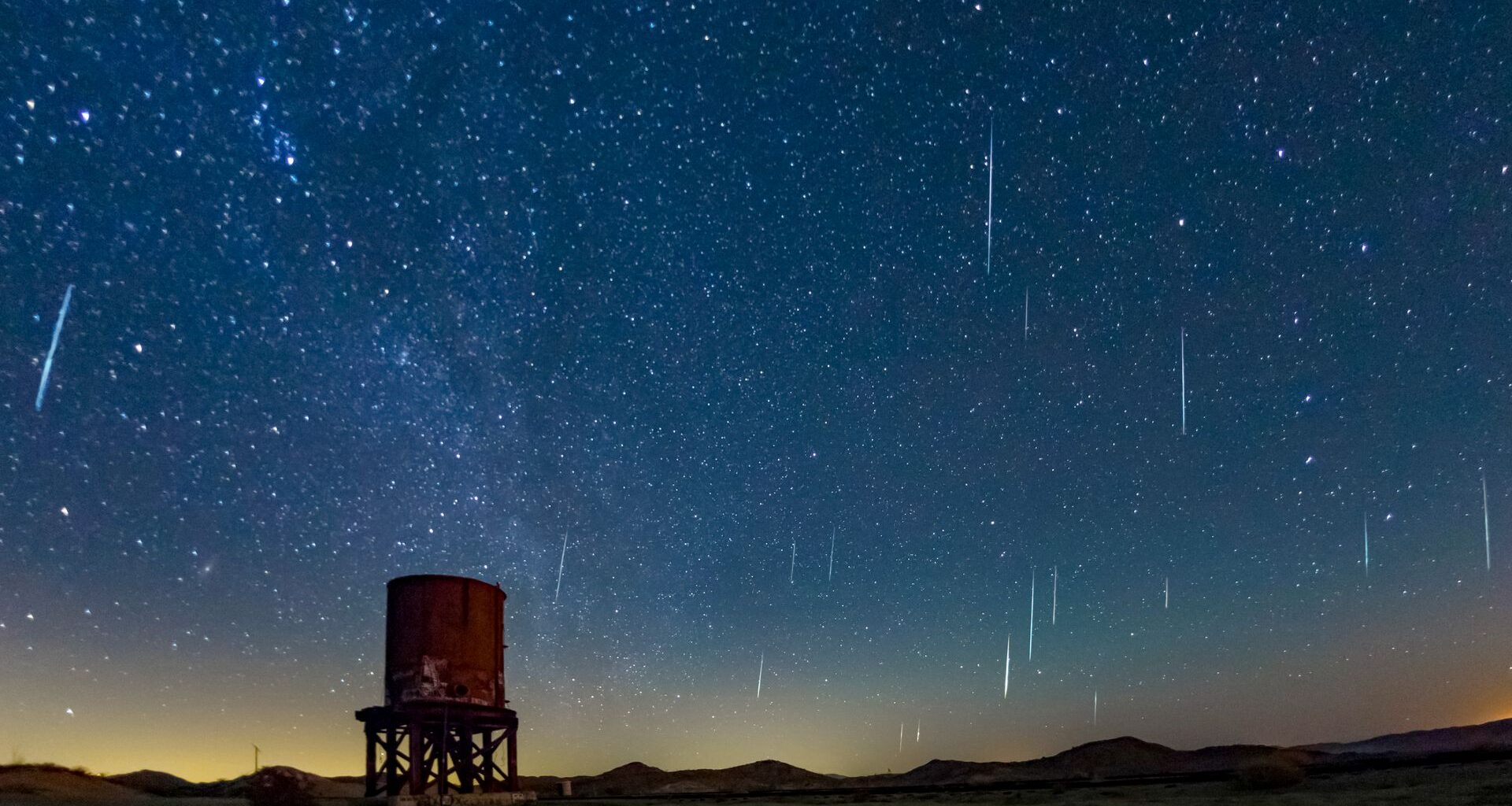 A dark blue night sky is streaked with white meteors moving through space with a rusty cylindrical water tower below.