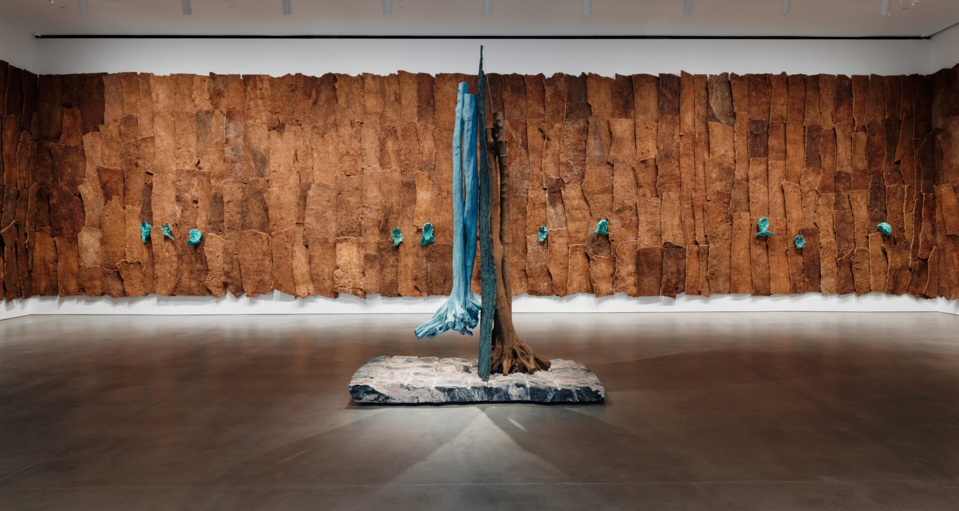 An installation view of Giuseppe Penone’s exhibition featuring a monumental wall covered in bark-like brown panels punctuated by turquoise sculptural forms, with a tree-like bronze sculpture suspended over a stone base in the center of the gallery.