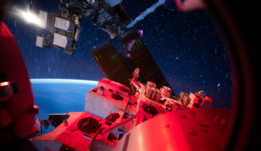 View from the International Space Station showing its illuminated exterior hardware in red light against a backdrop of deep space. Star trails form circular streaks across the sky due to the station’s motion, while Earth’s blue curvature glows softly at the horizon.