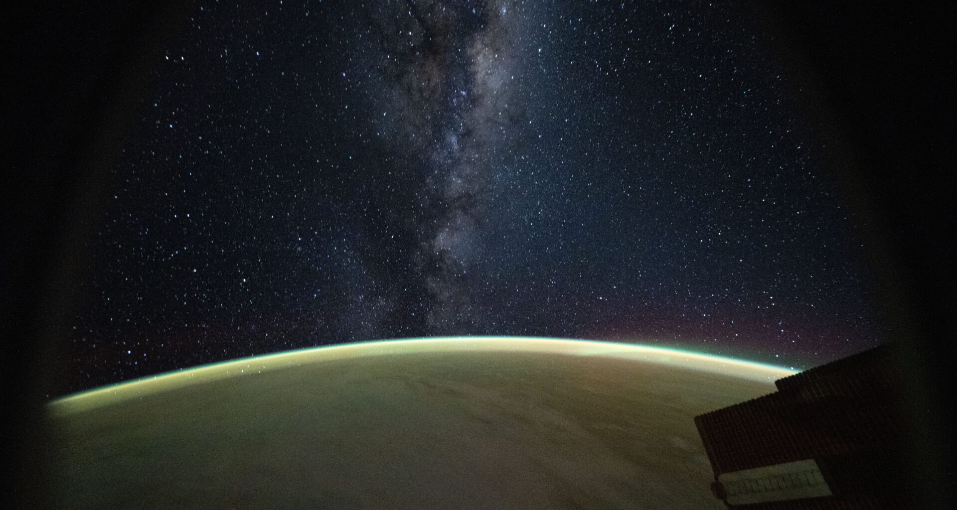 The Milky Way galaxy appears above Earth's greenish atmospheric glow. The galaxy's dust and stars can be seen against the darkness of space.