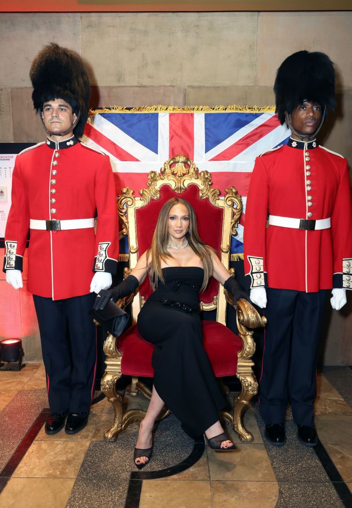 Jennifer Lopez sitting on a red and gold throne, flanked by two men in British Guard uniforms, in front of a Union Jack flag.