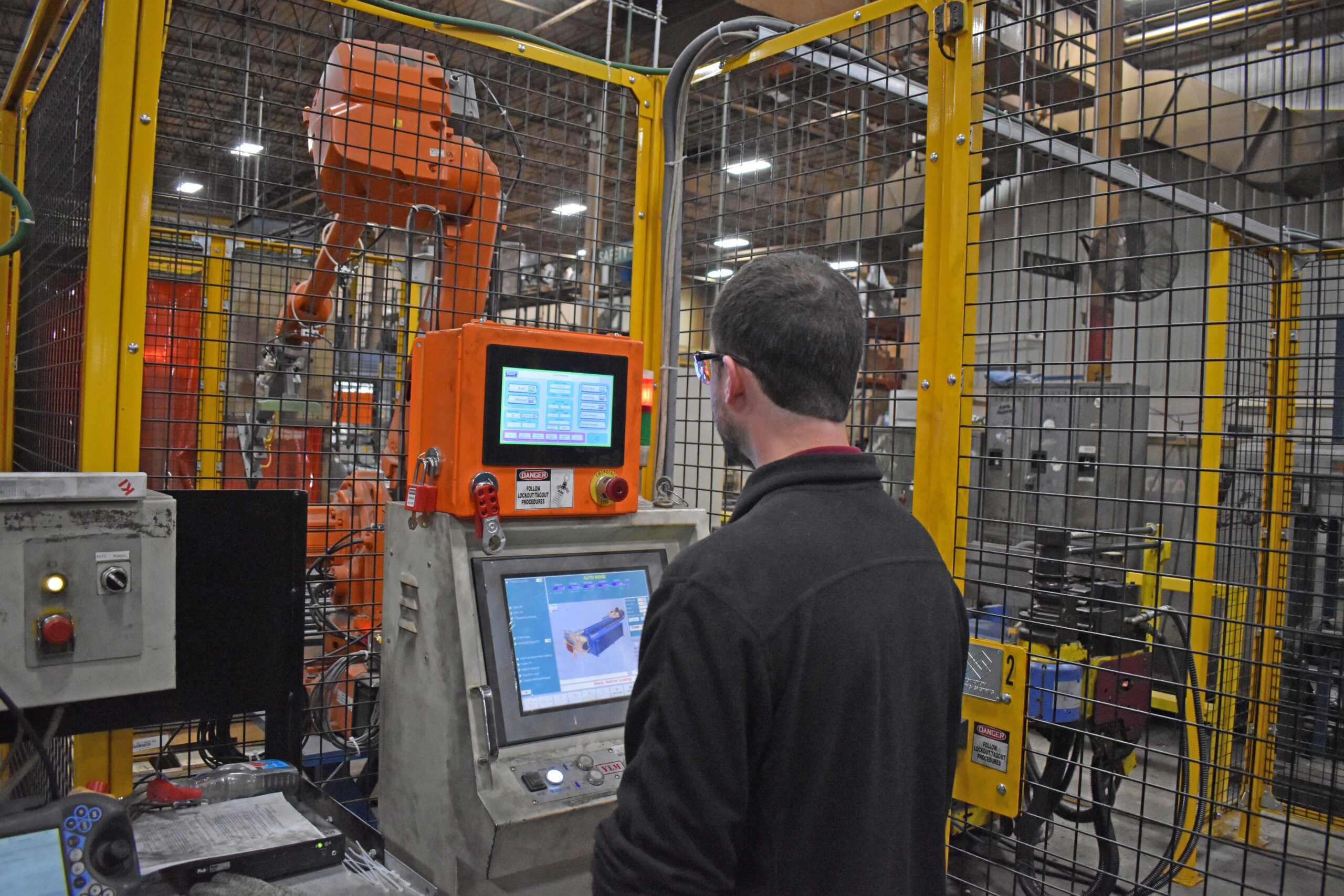 A person operates a computerized control panel in a factory setting with a robotic arm behind a safety cage.