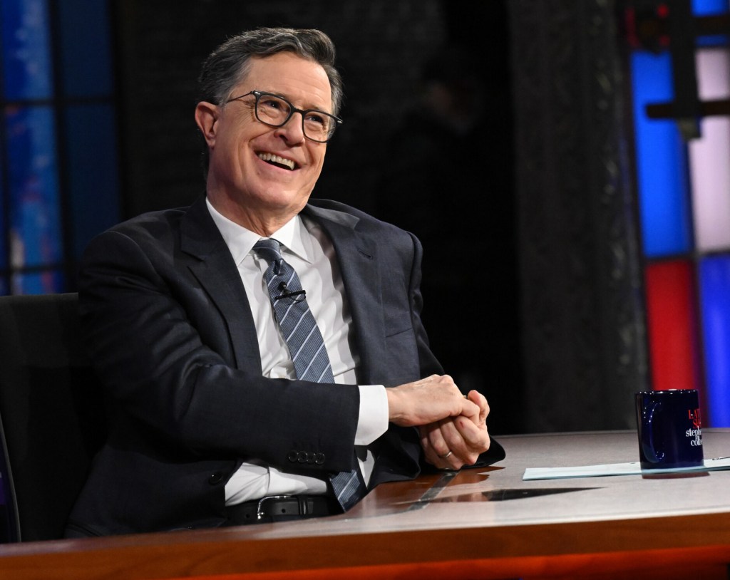 Stephen Colbert laughing while sitting at a desk with a mug.