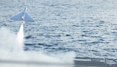 A military drone takes off from the flight deck of a combat ship. A rectangular satellite dish can be seen on the drone's top side.