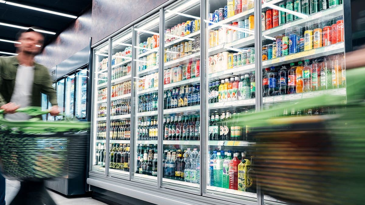 A man pushes a shopping cart in a grocery store in front of a selection of soft drinks.