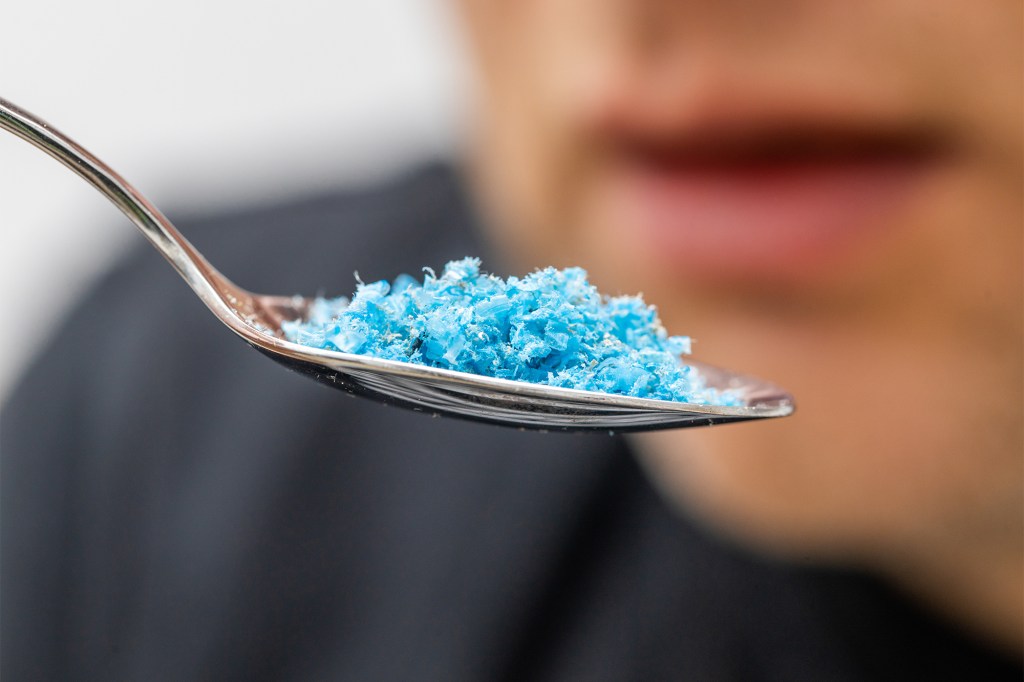 A person holding a spoon full of blue microplastic particles up to their mouth.