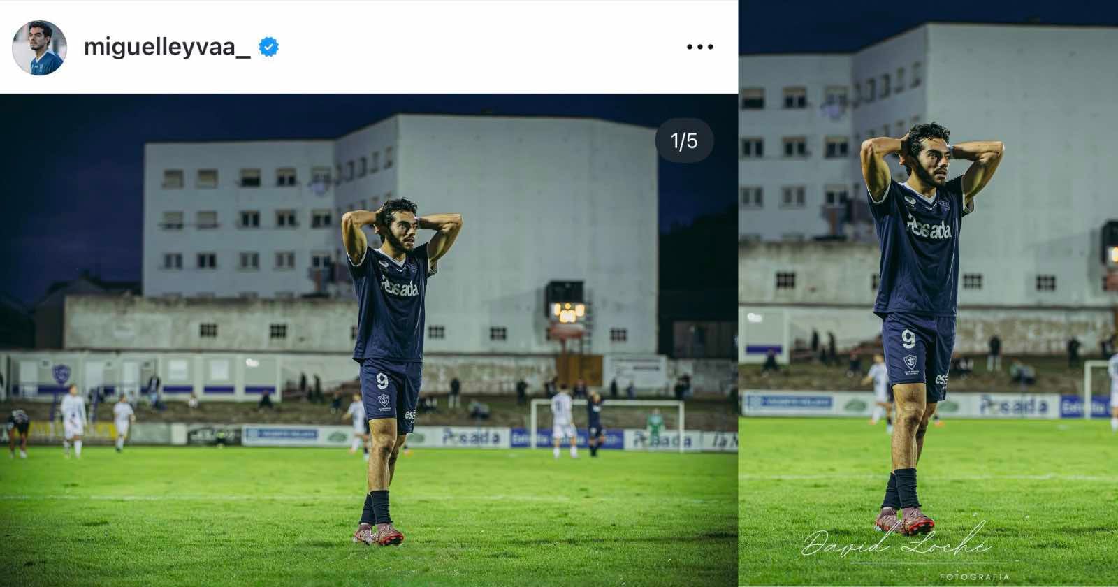 A soccer player wearing a navy blue jersey with the number 9 stands on a field with his hands behind his head during an evening match. Buildings and other players are visible in the background.