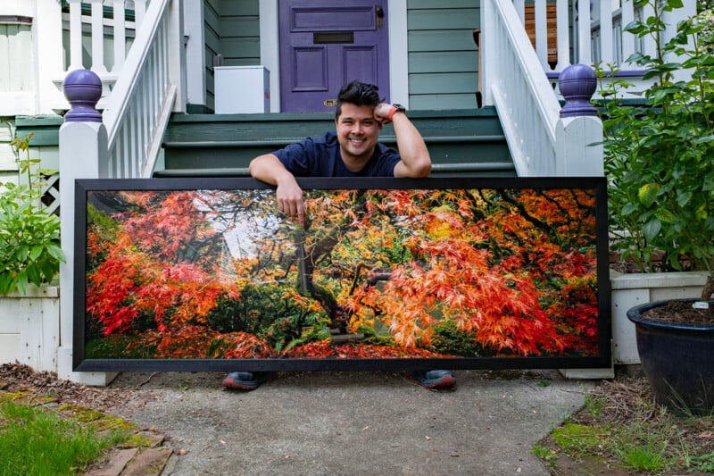 A person sits on outdoor steps, smiling and leaning on a large framed photograph of a vibrant autumn tree with red, orange, and yellow leaves. The house behind features white railings and a purple door.