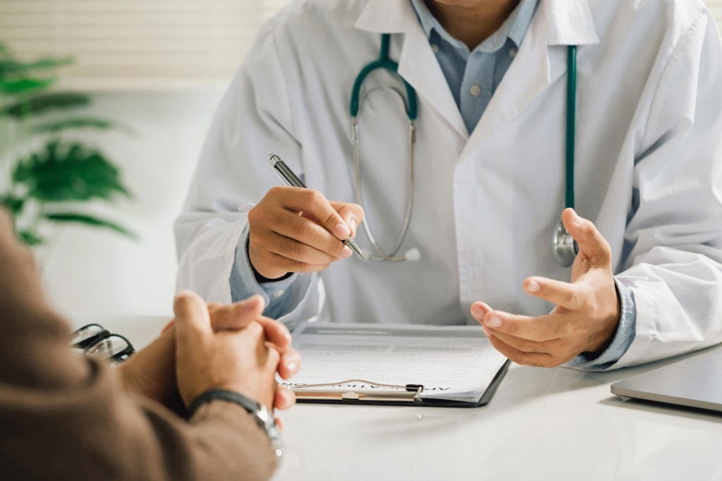 A doctor, in a white coat with a stethoscope, talks to a patient and holds a pen over medical papers on a clipboard.