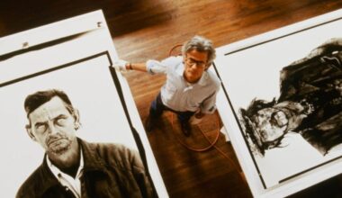 A man with glasses stands between two large black-and-white portrait artworks on the floor, looking up at the camera. The floor is wooden, and an orange cord is visible beside him.