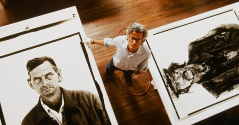 A man with glasses stands between two large black-and-white portrait artworks on the floor, looking up at the camera. The floor is wooden, and an orange cord is visible beside him.