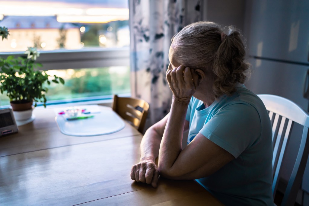 A sad, lonely older woman sitting at a table looking out a window.