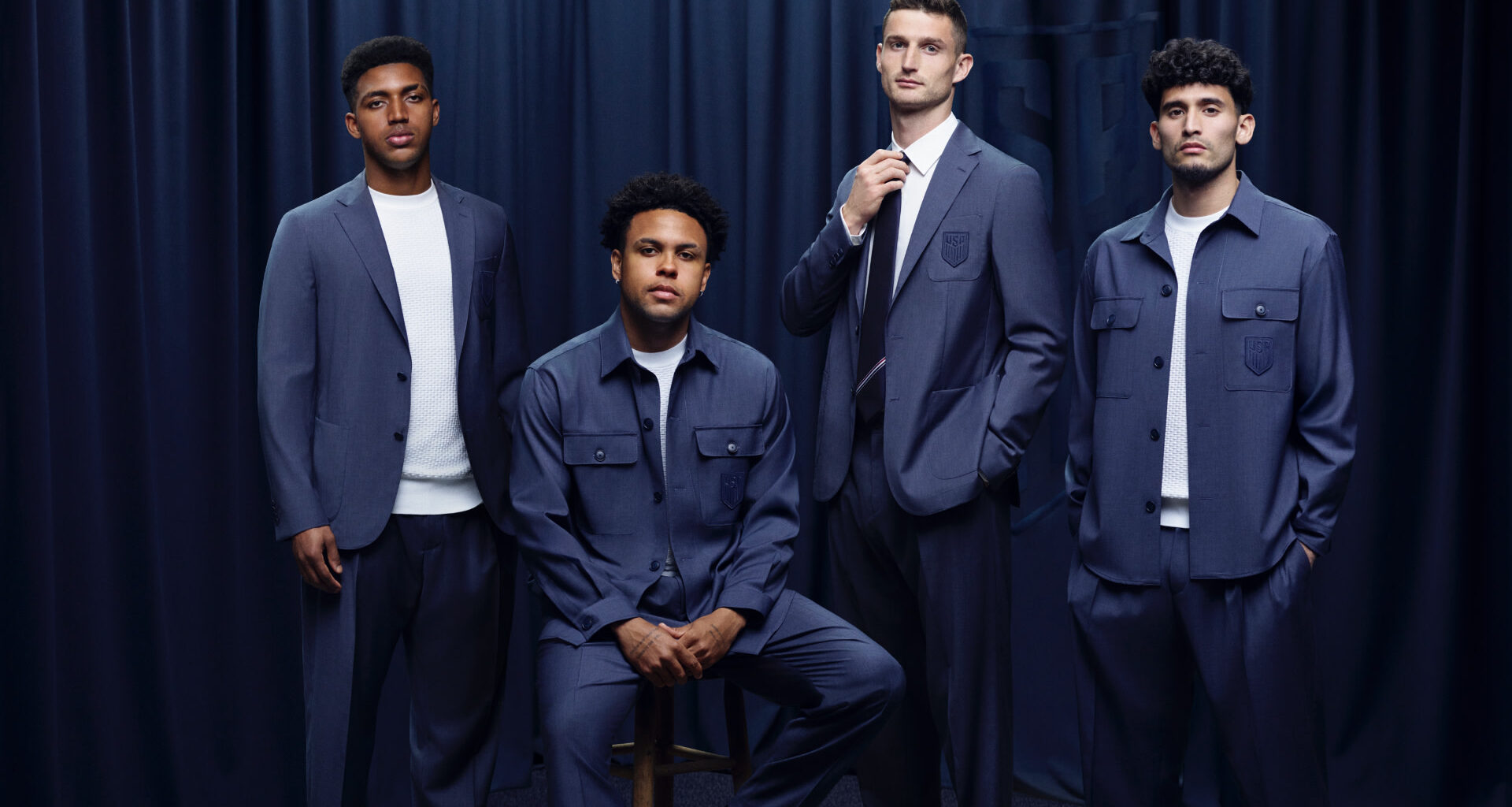 Four soccer stars in blue suits pose against a dark curtain backdrop. One is seated, while the others stand confidently beside him.