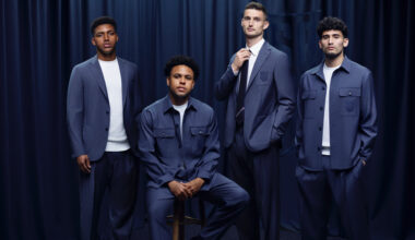 Four soccer stars in blue suits pose against a dark curtain backdrop. One is seated, while the others stand confidently beside him.