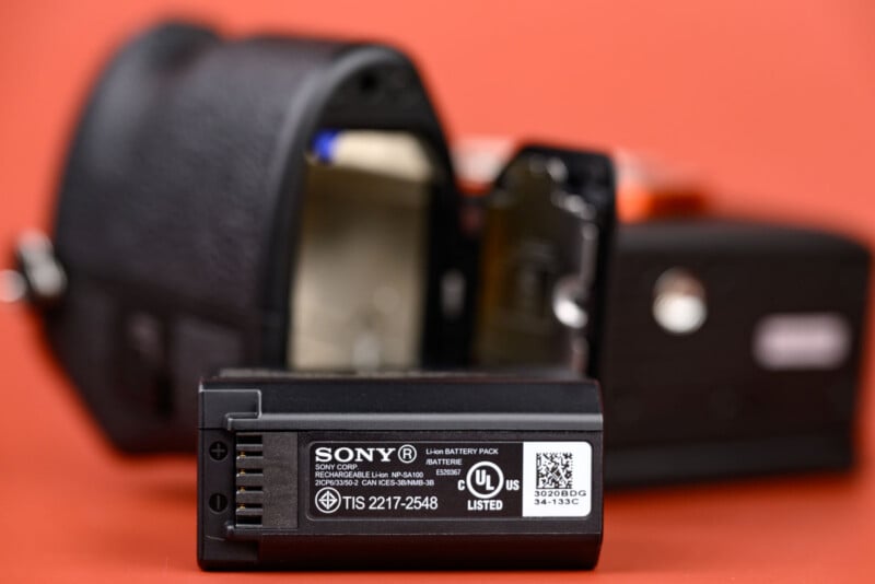 A close-up of a Sony camera battery in front of a blurred digital camera with an open battery compartment, set against a solid orange background.