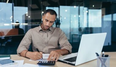 A person is using a calculator while sitting in front of a laptop in an office.