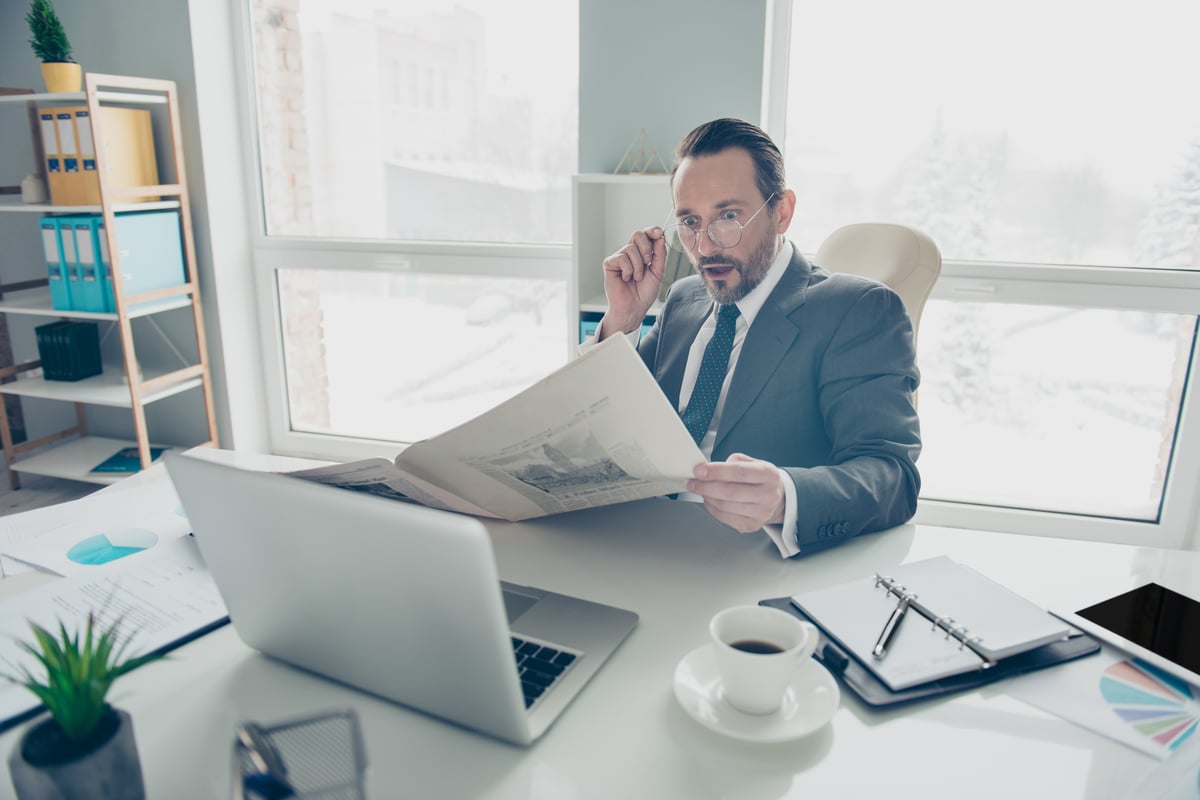 A slightly surprised-looking person reads a newspaper while sitting in an office.