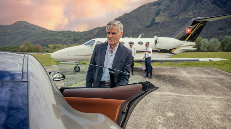 A mature man in a suit entering the passenger side of a luxury vehicle in front of a private jet.