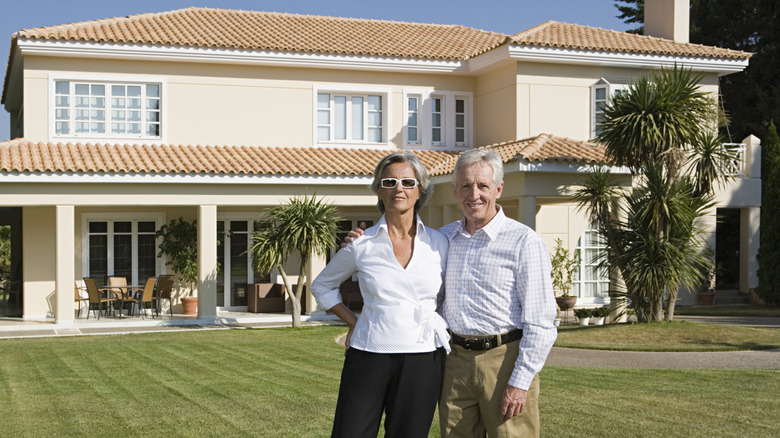 A wealthy retiree couple posing in front of a beautiful mansion.