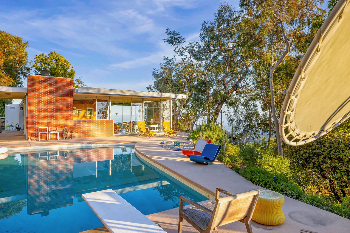 Modern house with large windows and patio beside a pool, surrounded by trees and outdoor furniture under a blue sky.