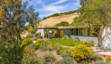 Mid-century modern house with large windows, surrounded by trees and hills under a clear blue sky.